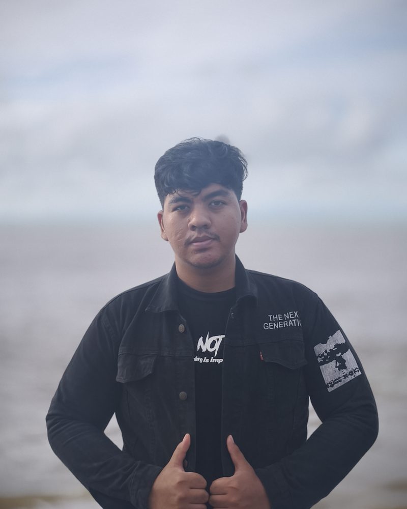 Aziz Nur Ashidiq standing on a beach in a black denim jacket, looking at the camera.